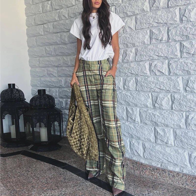 Woman wearing a white top and green plaid pants standing against a stone wall.