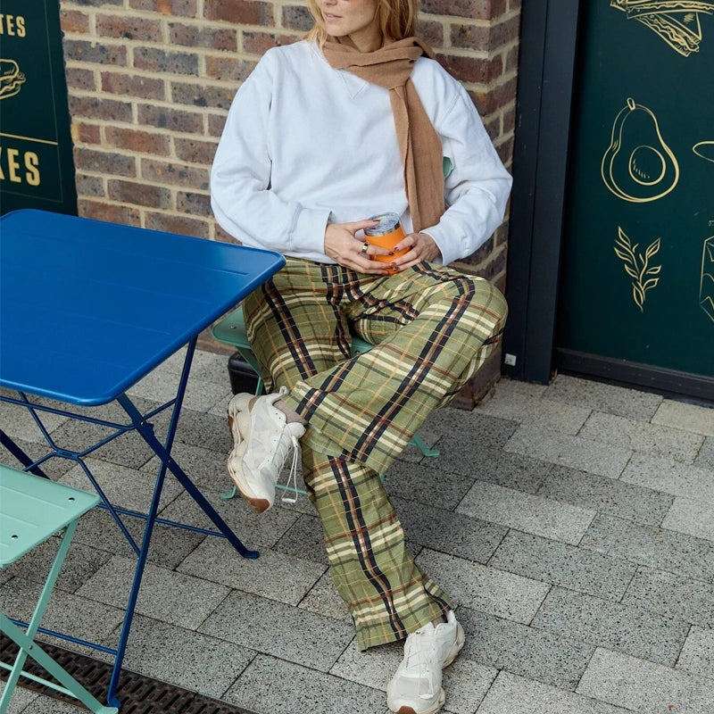 Person sitting outdoors holding a drink, wearing a white sweater and plaid pants, with a brick wall and green table in the background.