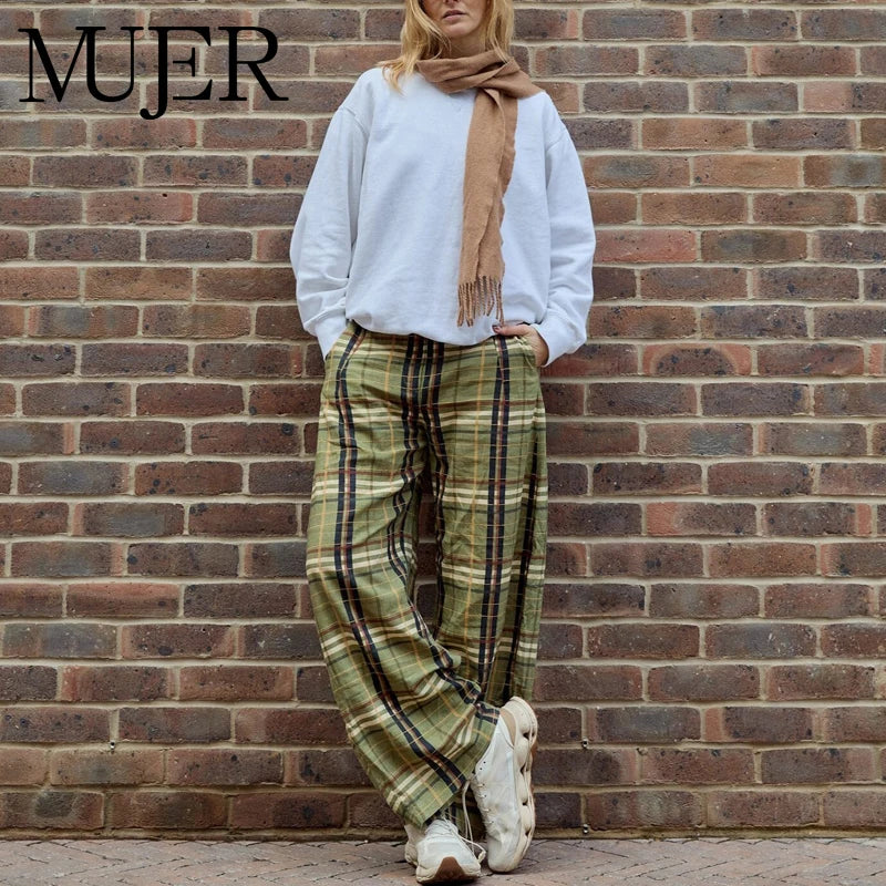 Person wearing a white sweater, plaid pants, and a brown scarf against a brick wall with 'MUJER' branding.